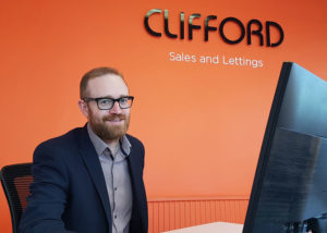 A man with a beard and glasses, wearing a suit, is smiling while sitting at a desk in front of a computer monitor. The background features an orange wall with the text "CLIFFORD Sales and Lettings" in black letters, hinting at the efficient management even for spaces like the storage room. - a room in the garden