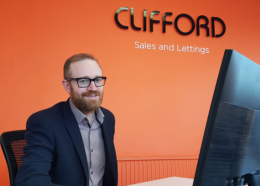 A man with a beard and glasses, wearing a suit, is smiling while sitting at a desk in front of a computer monitor. The background features an orange wall with the text "CLIFFORD Sales and Lettings" in black letters, hinting at the efficient management even for spaces like the storage room. - a room in the garden