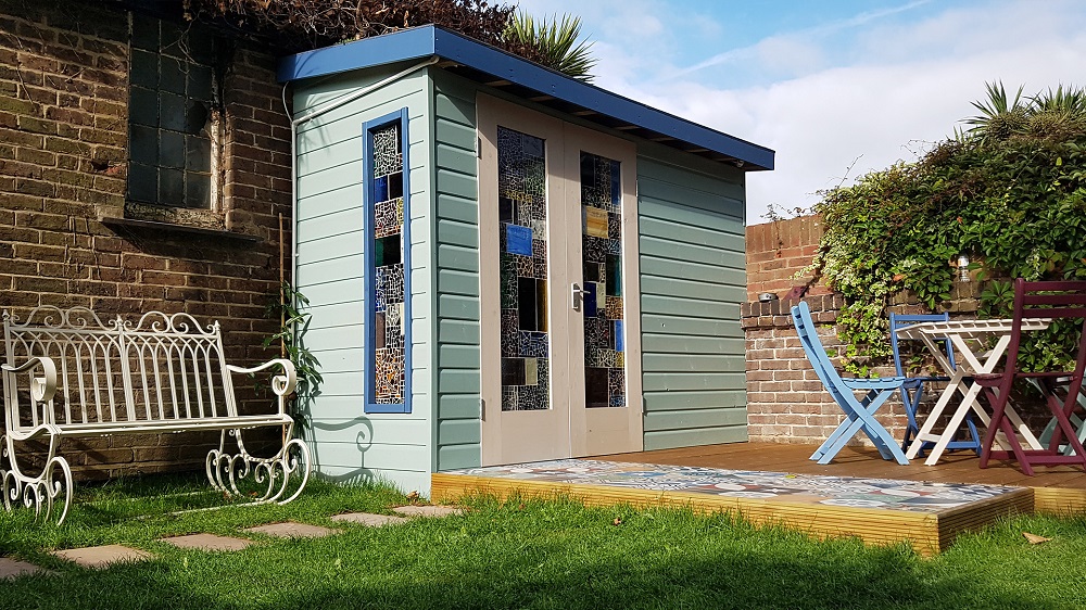 A quaint backyard scene with a small, light blue shed repurposed as a storage room, featuring colorful stained glass windows and a white door. In front of the shed is a tiled wooden deck, a white metal bench on the grass, and a purple and white patio set on the right. Greenery surrounds the area. - a room in the garden