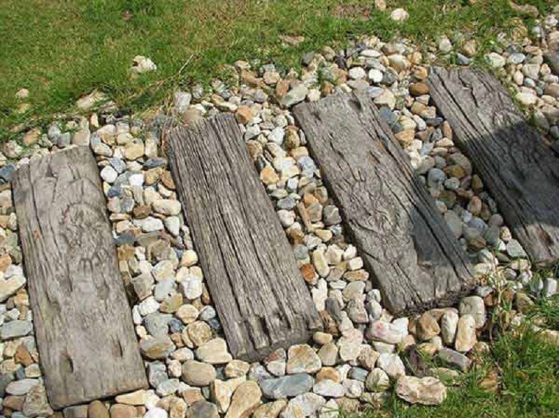 A garden path made of weathered wooden planks is set among a bed of small, multicolored pebbles. Green grass surrounds the pathway, adding a contrasting natural element. For charming and rustic path ideas, this blend of materials creates an inviting and picturesque scene. - a room in the garden