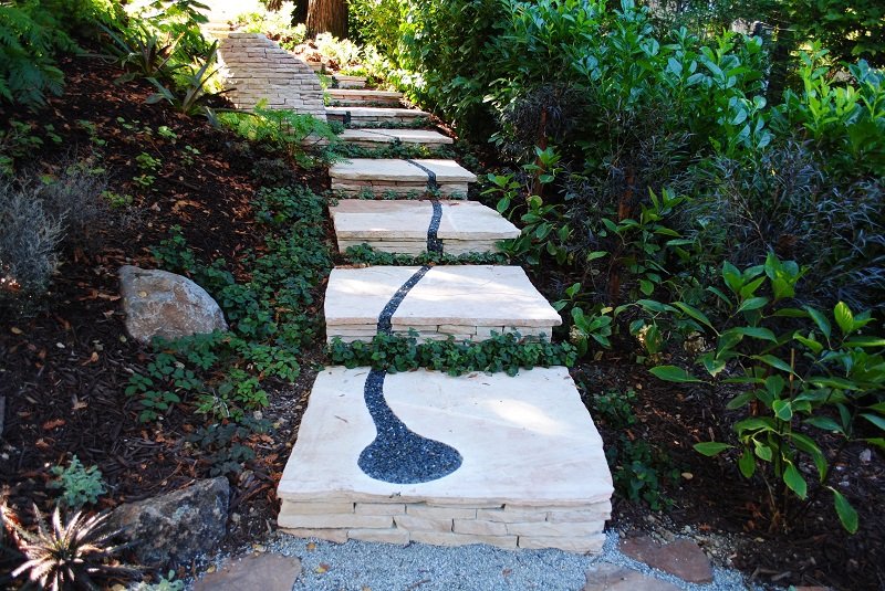 A garden path features large square stone slabs with a decorative black gravel inlay curving down the center. The stones are set among lush greenery and plants, creating a natural and tranquil atmosphere. For more path ideas, the steps continue up a gentle incline, leading out of view. - a room in the garden