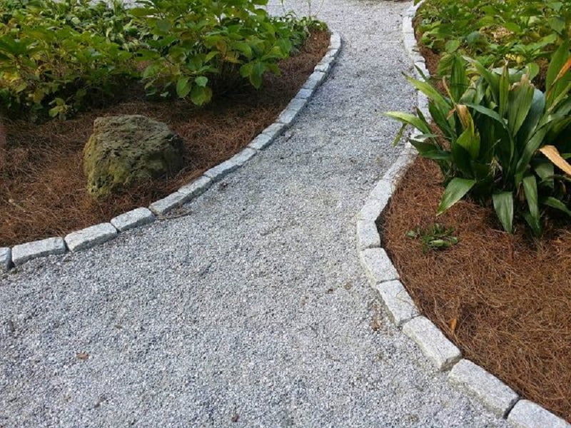 A gravel pathway curves through a garden, bordered by neatly arranged grey stones. The path is flanked by patches of green plants and mulch, with a large rock visible on the left side among the vegetation, offering inspiring path ideas for serene landscapes. - a room in the garden