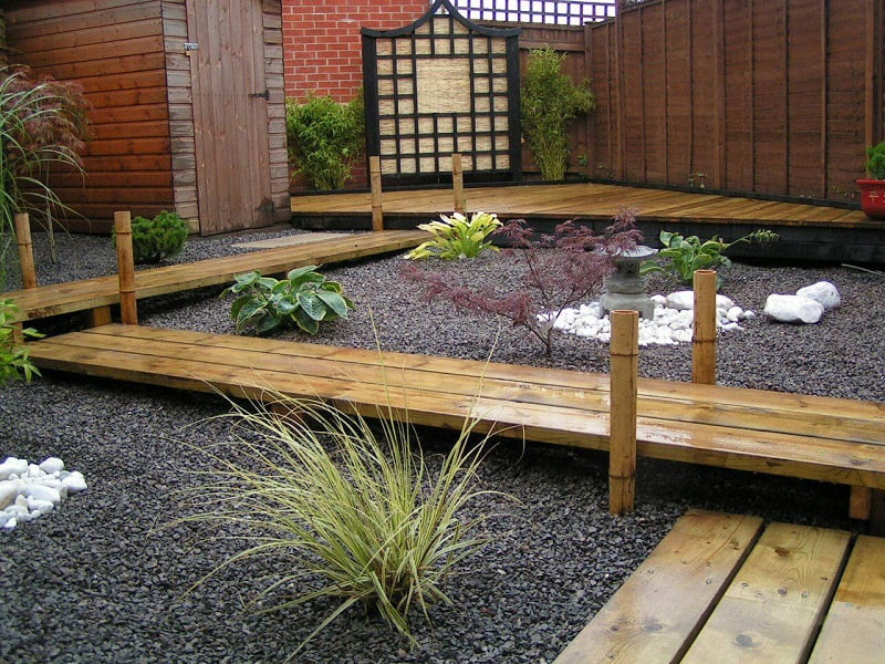 A minimalist Zen garden features wooden walkways over dark gravel, various green plants, and a Japanese maple tree. A wooden fence and shed are in the background. The layout, with its serene path ideas, creates a balanced atmosphere. - a room in the garden