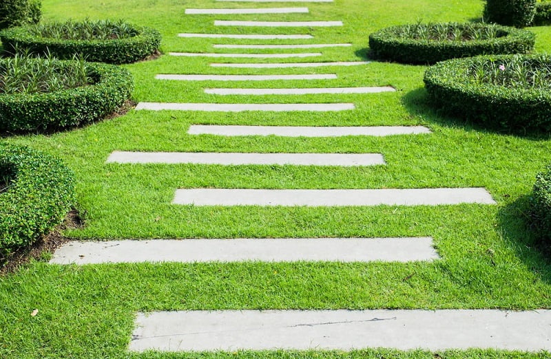 A neatly manicured garden path, ideal for those seeking path ideas, consists of rectangular concrete slabs set into a bright green lawn. The path is bordered by circular and rectangular-shaped hedges, adding a structured, geometric aesthetic to the garden. - a room in the garden