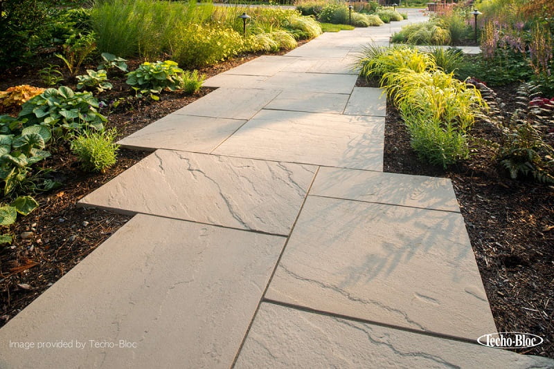 A neatly landscaped garden path, ideal for those seeking path ideas, is paved with large, beige stone tiles bordered by various green plants and shrubs. The path curves gently in the distance, surrounded by well-maintained greenery. The image includes a small Techno-Bloc logo in the bottom right corner. - a room in the garden