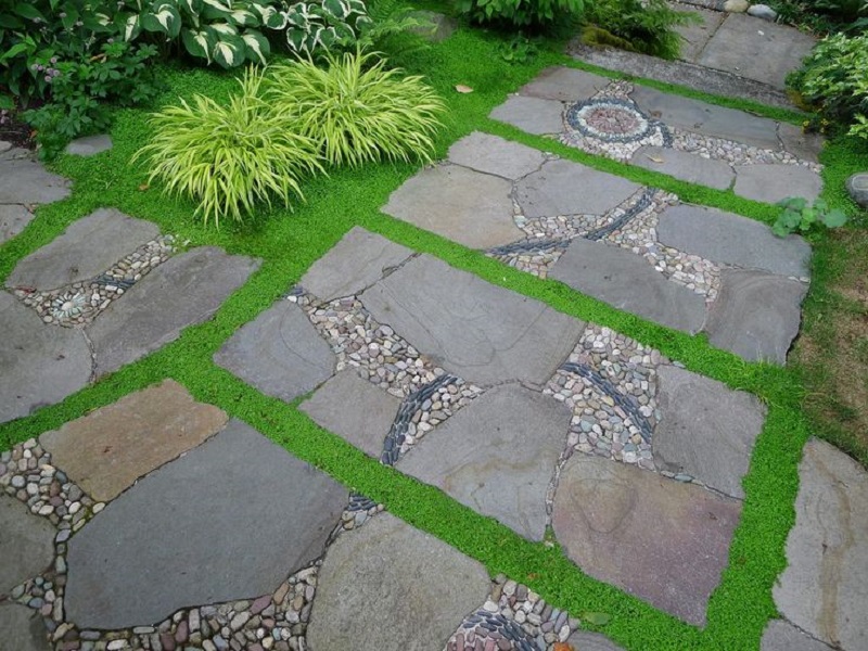 A beautifully designed stone pathway featuring large, irregularly shaped stones set in a grid pattern, bordered and interspersed with small pebbles and green moss. Perfect for those seeking path ideas, the lush greenery and ornamental grass enhance the natural look of the path. - a room in the garden