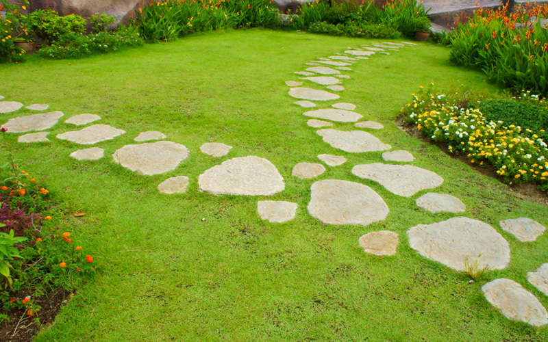 A well-maintained garden with a lush green lawn features a winding stone path, showcasing creative path ideas as it leads through the space. The path is bordered by a variety of colorful flowers and green plants, creating a vibrant and serene atmosphere. - a room in the garden