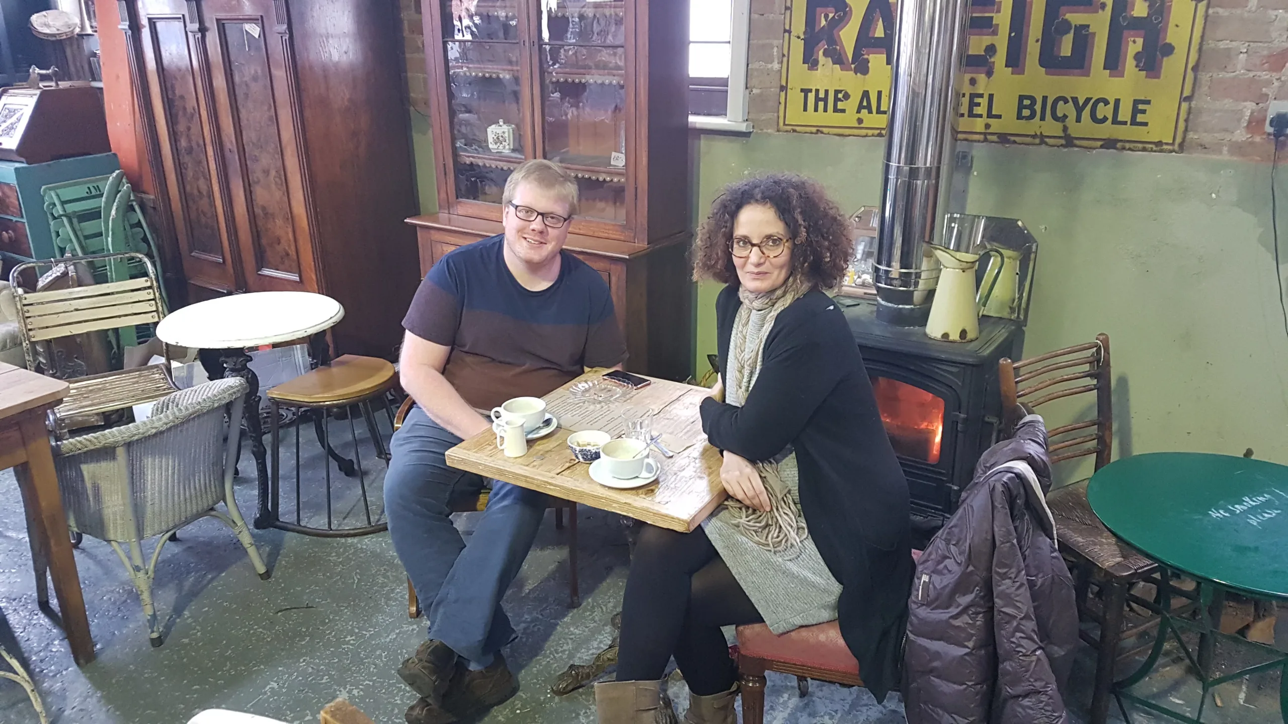 Two people are sitting at a small wooden table in a cozy cafe. The man is wearing glasses and a dark t-shirt, while the woman has curly hair and wears a grey dress with a black jacket. They are smiling, and there are cups and a teapot on the bespoke design table. A vintage "Raleigh" bicycle sign adorns the wall behind them. - a room in the garden