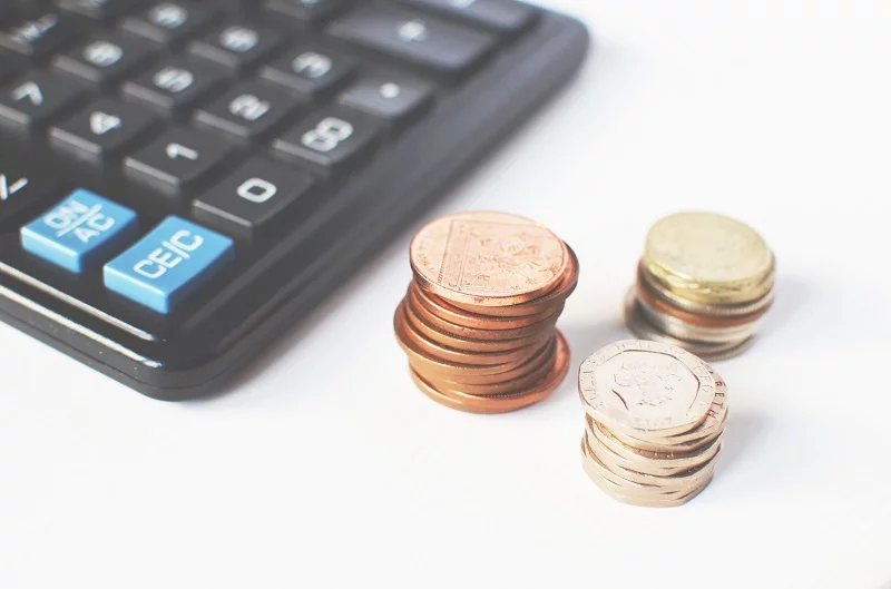 Close-up of a black calculator with blue and white keys, boasting a bespoke design, sits next to three stacks of coins on a white surface. The coins are of varying denominations and sizes. - a room in the garden