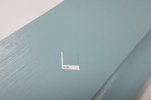 A small paint sample card with the designation "18C 53" is placed on top of a large wooden surface painted in a matching shade of light blue. The photo highlights the color comparison between the sample card and the wood paint on the surface. - a room in the garden