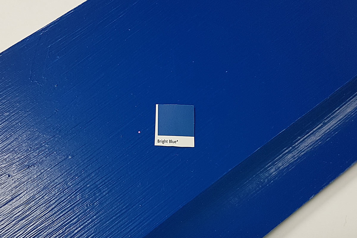 A sample paint swatch labeled "Bright Blue" is placed on top of a surface painted in vibrant blue wood paint. The image highlights the similarity between the swatch and the paint on the surface. - a room in the garden
