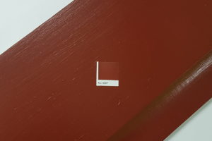 Sample of brown wood paint on a wooden surface, with a small label in the center reading "RAL 3009." The paint has a smooth finish, and there are light reflections showcasing its sheen. - a room in the garden