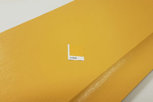 A sample card labeled "Sunshine" is placed on a wooden plank painted in a similar shade of yellow. The wood paint gives the plank a smooth, glossy finish. - a room in the garden