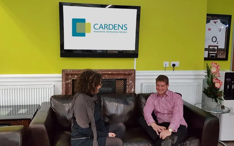 Two people sitting on a brown leather sofa, engaged in conversation. Behind them, a wall-mounted TV displays the "CARDENS Accountants and Business Advisors" logo. The room, akin to a garden office, features a lime green wall, framed sports jerseys, and a small table on the left. - a room in the garden