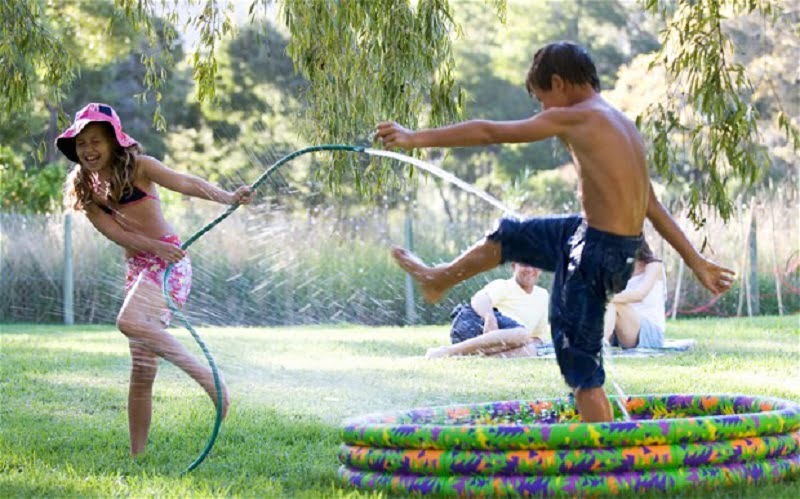 Two children play with a hose outside on a sunny day. A girl in a pink hat and swimsuit sprays water from the hose at a boy in swim trunks standing in an inflatable kiddie pool. The boy lifts one leg in the air while several people, dreaming of ways to connect water to my garden room, sit on the grass in the background. - a room in the garden