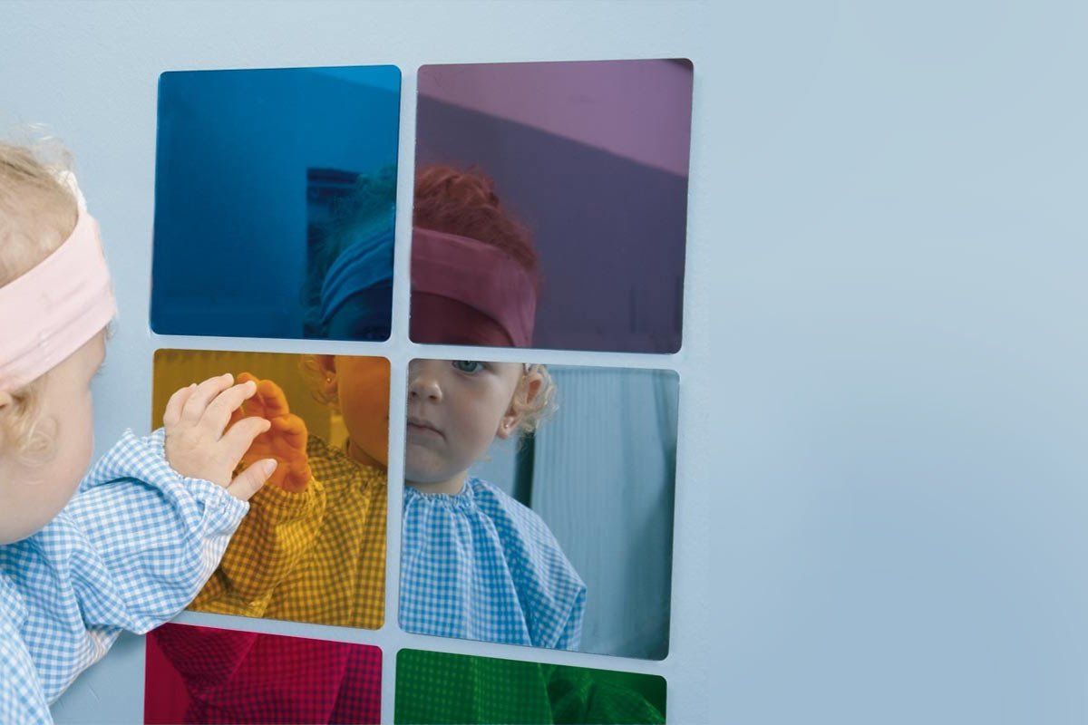 A toddler wearing a checkered bib and a headband touches a wall mirror made up of six small, colorful tiles in blue, purple, yellow, red, and green. The toddler's reflection is visible in the tiles against the light blue background—a perfect touch for garden baby room ideas. - a room in the garden