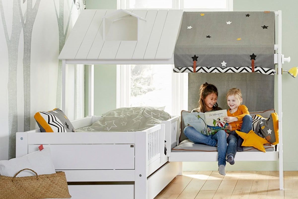 Two children sit together in a cozy, white house-shaped bed with a canopy adorned with stars. They are happily reading a book, surrounded by plush pillows, including one in the shape of a star. The room, inspired by Garden Baby Room Ideas, has light green walls with a tree mural and wooden flooring. - a room in the garden