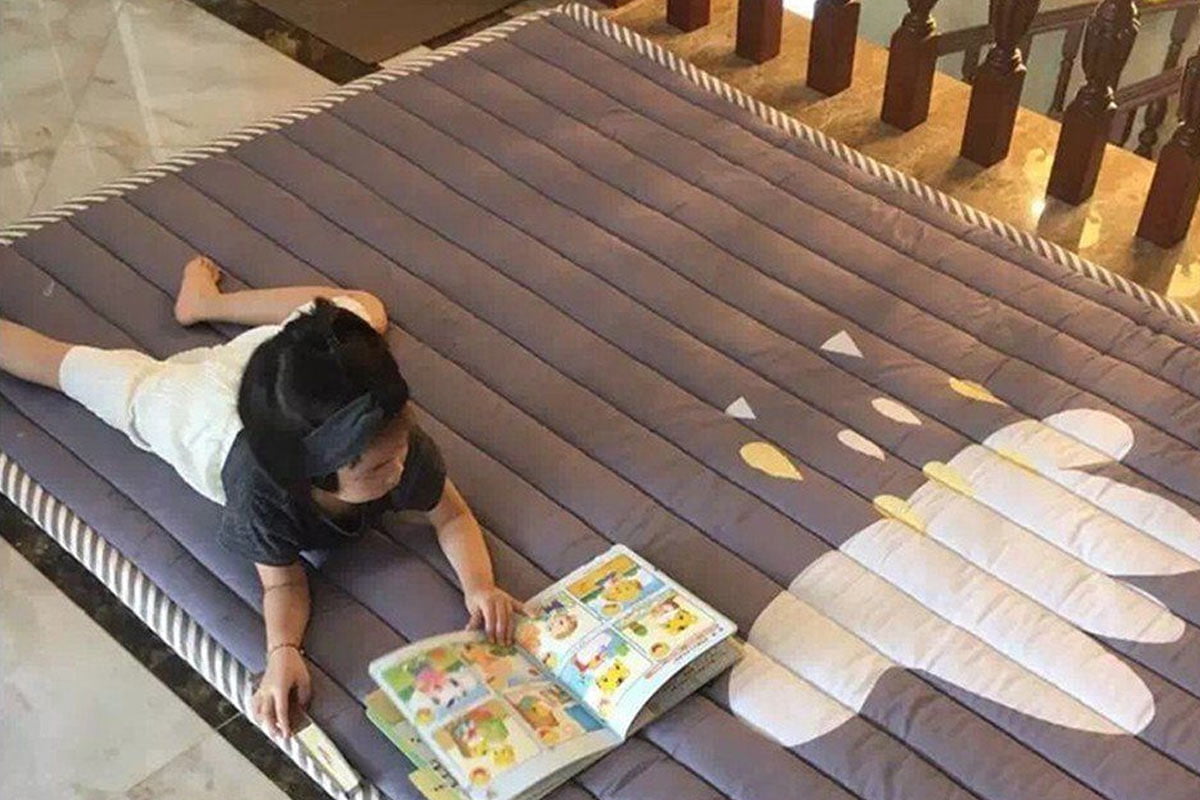 A young child is lying on their stomach on a large, grey, cushioned mat, looking at an open picture book. The mat features a white and yellow design, perfect for those seeking garden-themed baby room ideas. The child appears to be indoors, with wooden railings in the background. - a room in the garden