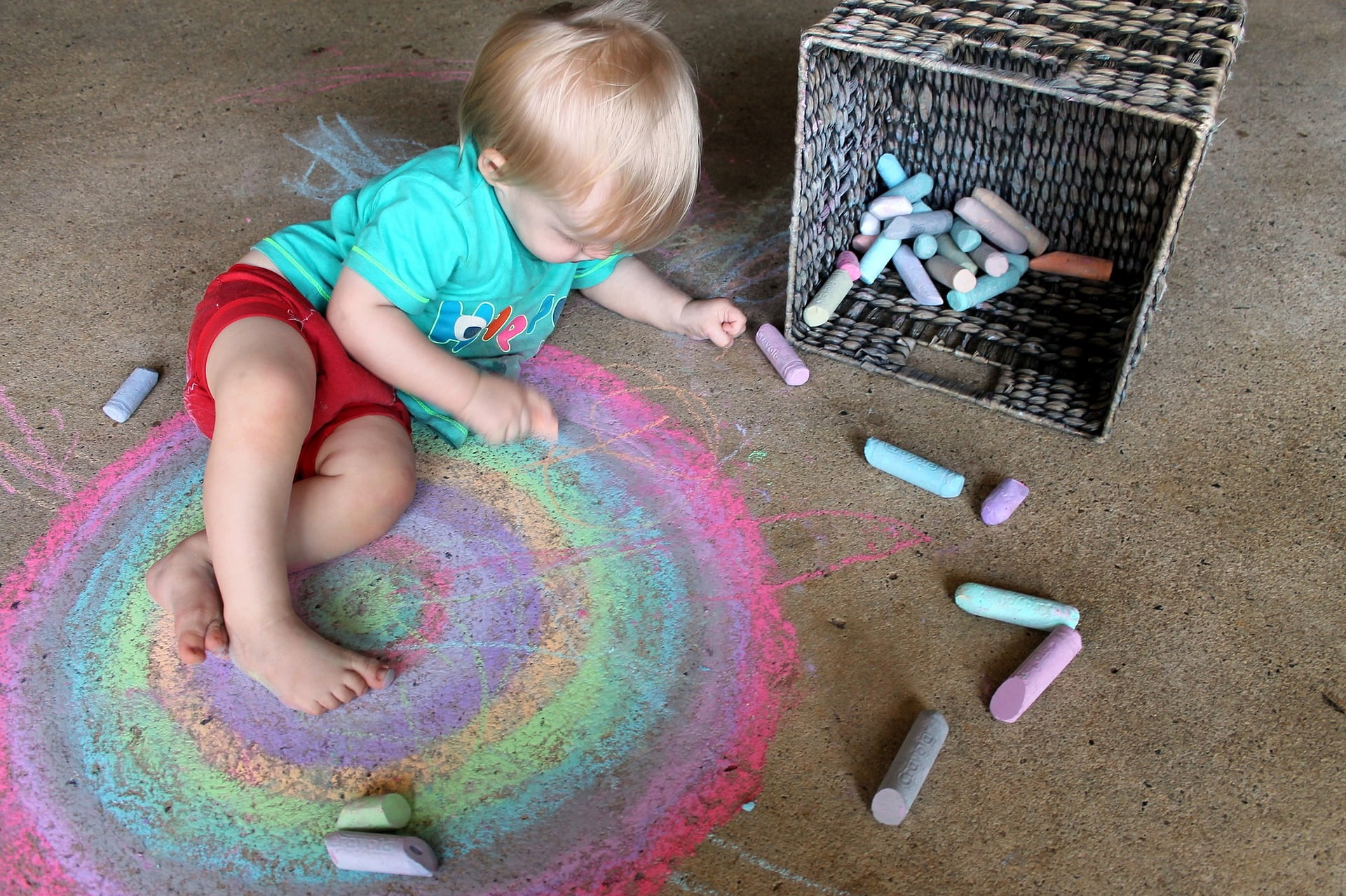 A toddler with light hair wearing a turquoise shirt and red shorts sits on the ground in the garden, drawing with colorful chalk. Around the child, there is a large multicolored chalk drawing, and a woven basket is tipped over, scattering more chalk pieces onto the ground. - a room in the garden