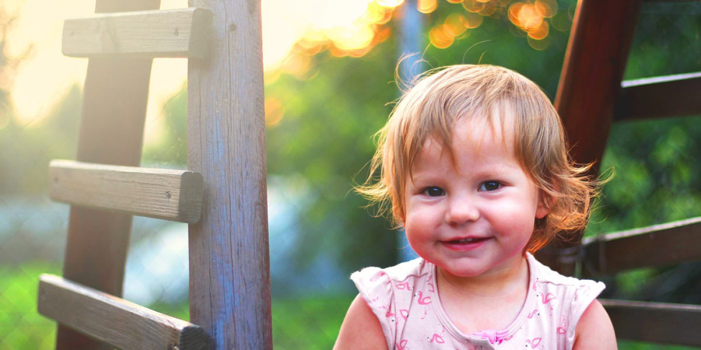 A smiling toddler with short, light brown hair and a pink dress stands beside a wooden ladder in a sunny garden. The background shows blurred greenery and a bright, warm light, creating a cheerful and vibrant atmosphere perfect for baby room ideas. - a room in the garden