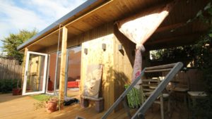 A wooden cabin with a slanted roof, featuring an outdoor area with a wooden deck. A partially rolled-up curtain hangs on the right side, revealing an outdoor dining table with chairs. A Buddha face sculpture leans against the cabin wall, showcasing garden buildings in their natural serenity. The sun is shining brightly. - a room in the garden