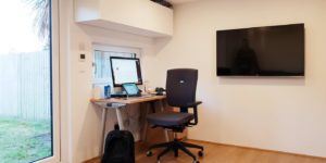 A minimalist garden office with a desk setup by a window. The desk has a computer monitor, keyboard, mouse, and various office supplies. A black office chair is in front of the desk, and a black backpack sits on the floor. A flat-screen TV is mounted on the wall. - a room in the garden