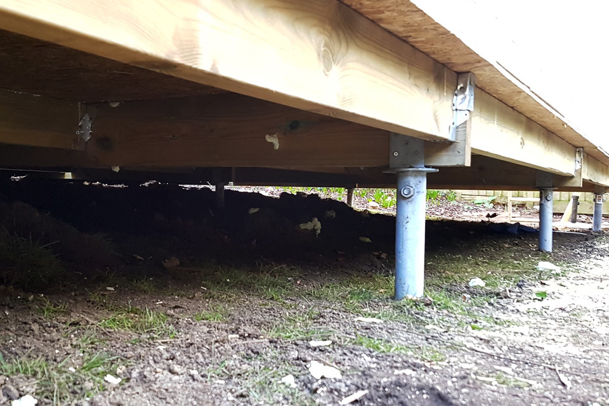 A close-up view of the underside of a wooden deck. The deck is elevated on metal support posts, forming sturdy foundations. The ground beneath is a mix of dirt and sparse grass. The wooden boards of the deck are secured with metal brackets, and some sunlight filters through the gaps. - a room in the garden