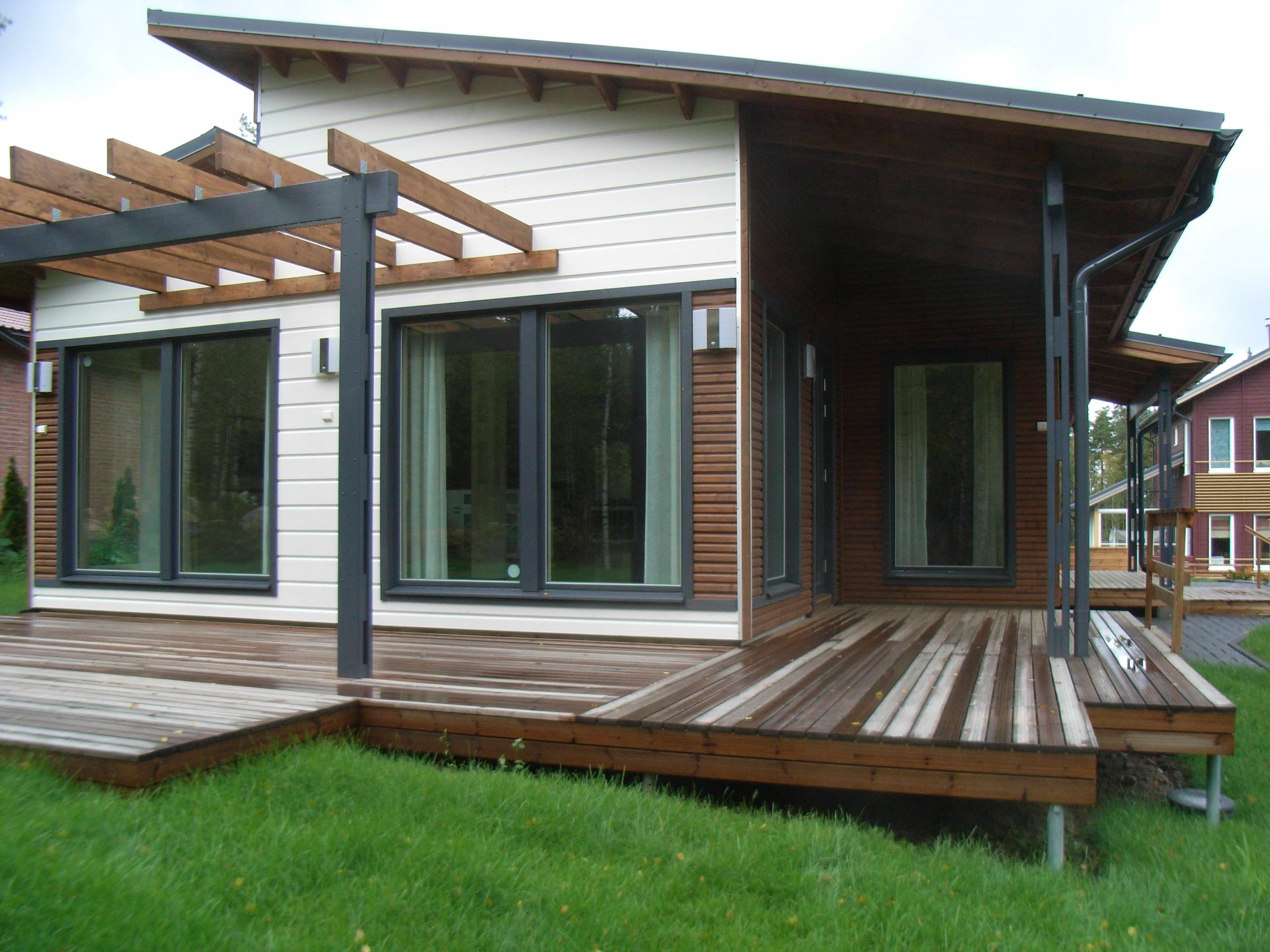 A modern house with a mix of white and brown wooden exterior features large windows and a wooden deck that wraps around the building. The deck is partially covered with a pergola, establishing solid foundations of style. The house is set on a grassy lawn with another similar house visible in the background. - a room in the garden