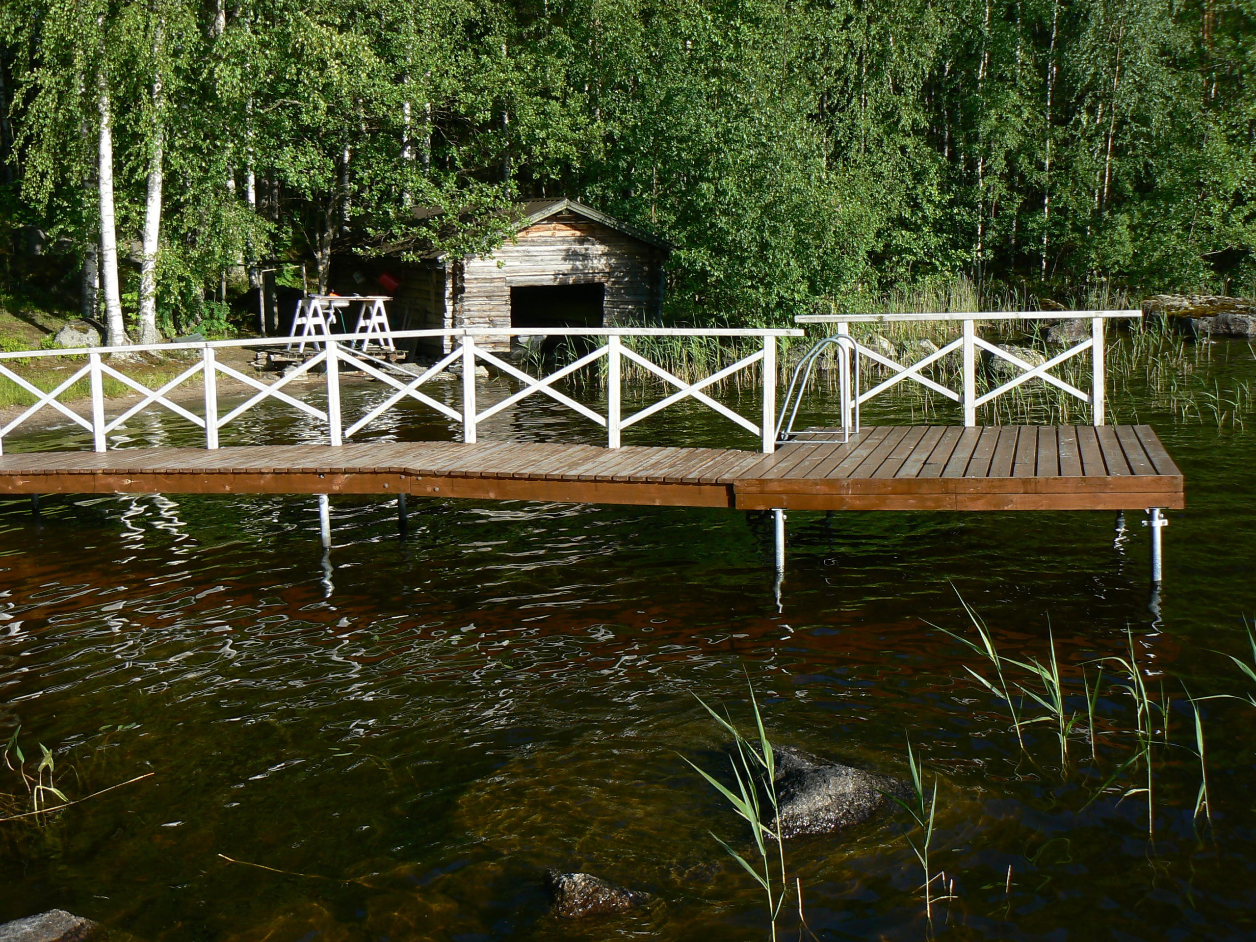 A wooden dock, with white railings and a ladder leading into the water, extends into a calm lake surrounded by lush greenery. In the background, there's a small wooden cabin nestled among trees, its foundations blending seamlessly with nature. - a room in the garden