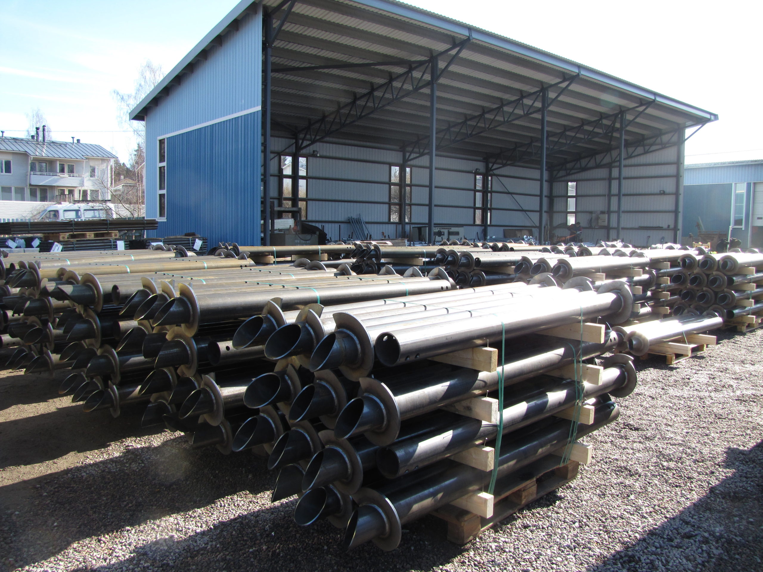 Stacks of large metal pipes are arranged on pallets outside a blue industrial building, providing strong foundations for future projects. The gravel-covered area features other structures and buildings visible in the background under a clear sky. - a room in the garden