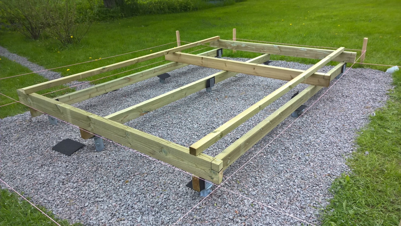 A wooden foundation frame for a small structure is being constructed on a gravel bed. Elevated on metal brackets, the foundations are outlined by strings marking the boundaries around the structure. The surrounding area is grassy. - a room in the garden