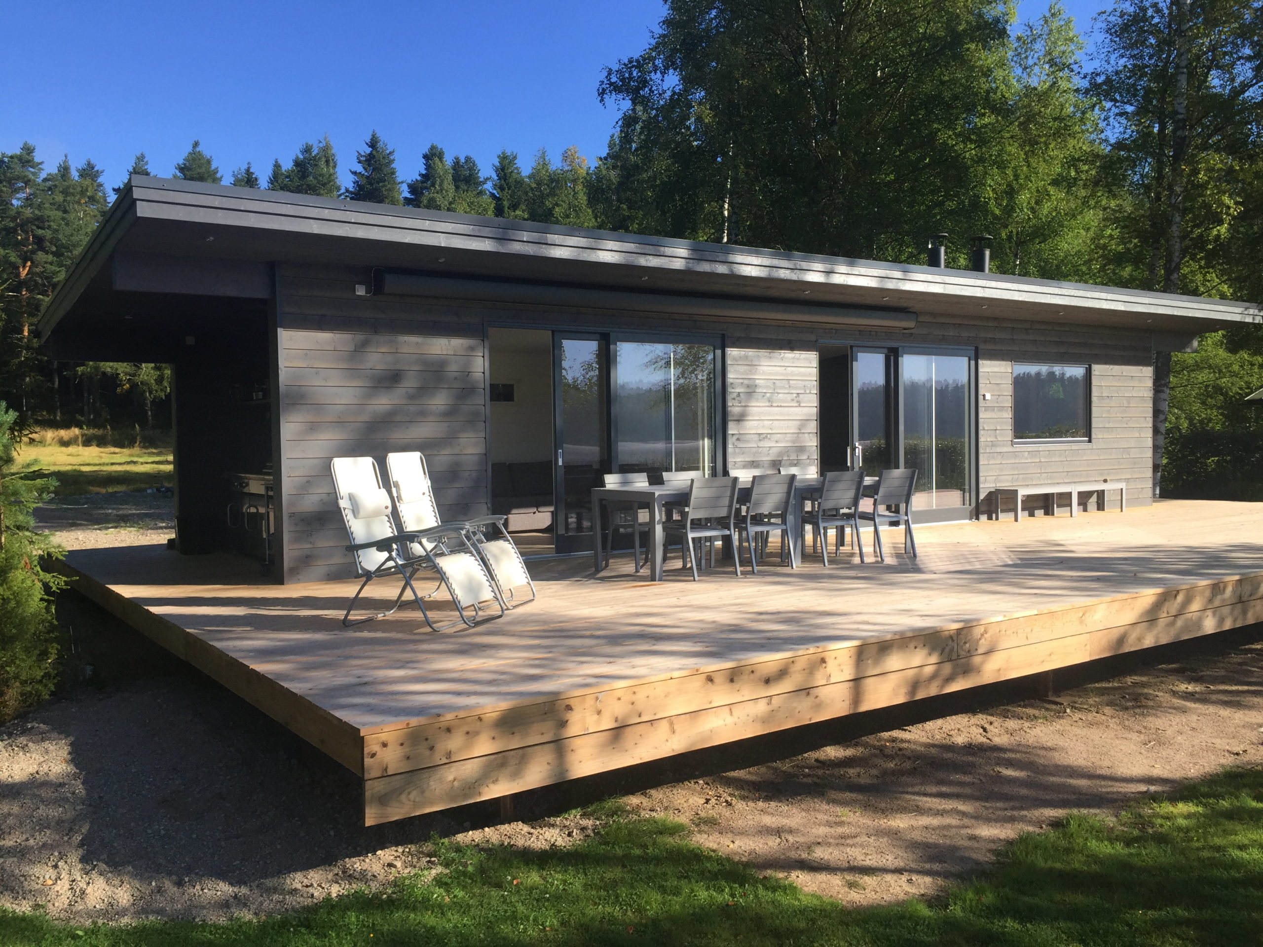 A modern, single-story cabin with dark wood paneling sits in a sunny, wooded area. Built on sturdy foundations, the house features large glass sliding doors and a spacious wooden deck with patio furniture and two lounge chairs, surrounded by green grass and tall trees. - a room in the garden