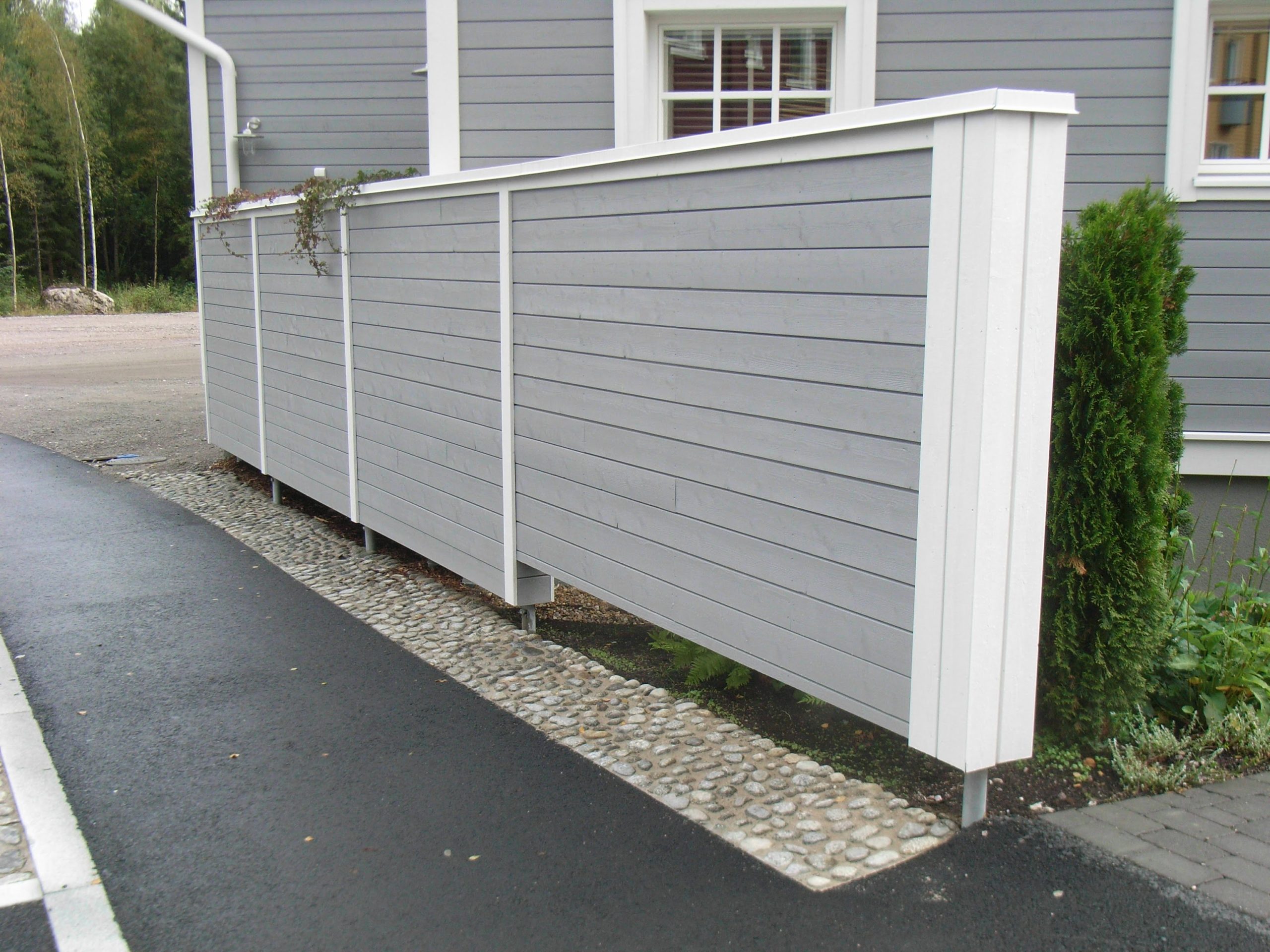 A modern gray and white wooden fence stands next to a gray house with white trim, elevated on metal pedestals that serve as its foundations. The fence is set on a gravel path bordering a paved road, with a few green shrubs and a window visible near the house. - a room in the garden