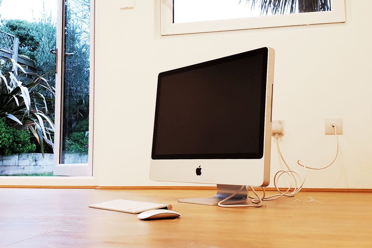 A silver iMac computer with a black screen is placed on a wooden floor. A keyboard and mouse lie nearby. The computer is plugged into a wall outlet with a cable trailing to the right. A glass door to the left opens to reveal the garden building visible in the background. - a room in the garden