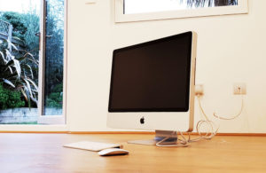 A desktop computer with a black screen, keyboard, and mouse sits on a wooden floor near a white wall with a power outlet. The room features a glass door with a view of the garden building and outside greenery, including trees. - a room in the garden