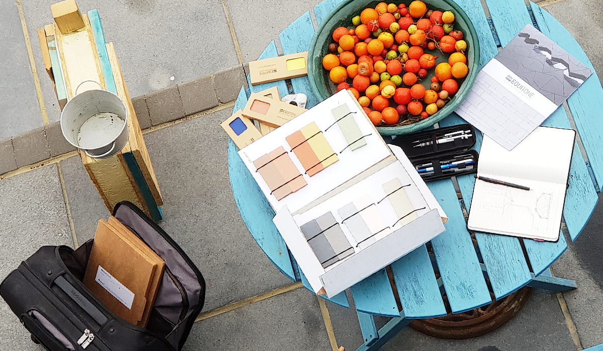 A bright outdoor scene includes a turquoise wooden table with a bowl of cherry tomatoes and various items such as color swatches, a notepad with sketches, pens, and a magazine. The setting evokes the creative vibe of designer garden homes, while nearby, a black backpack with notebooks peeks into the frame. - a room in the garden