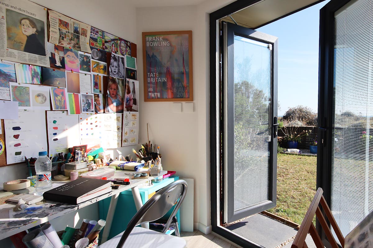 A bright, sunlit art studio with an open door leading outside. The workspace resembles a garden room, featuring a cluttered desk with art supplies, brushes, and paint. The walls are adorned with various artworks and posters. A chair is pulled up to the desk. The outdoor area is grassy. - a room in the garden