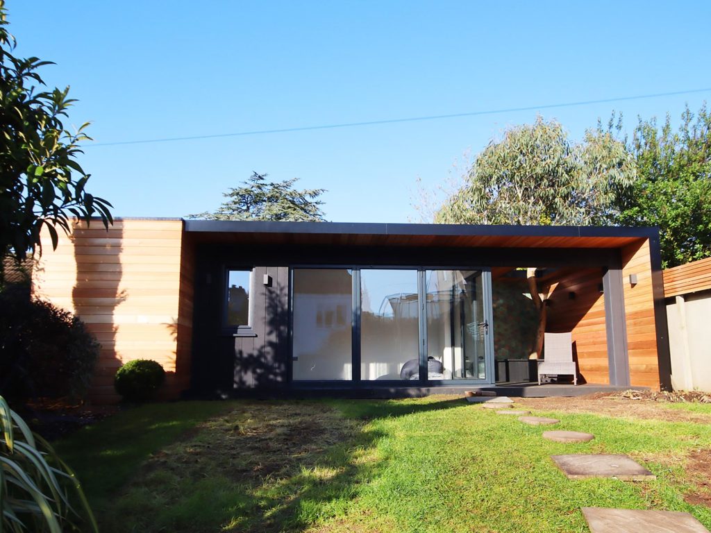 A modern, single-story house featuring a facade with large glass sliding doors and wooden paneling. Surrounded by a neatly maintained lawn with stepping stones leading to the entrance, the home boasts designer garden rooms. The sky is clear and blue, while trees and shrubs frame the scene. - a room in the garden