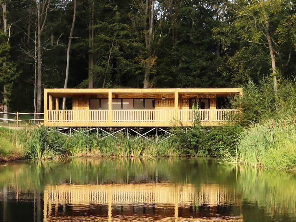 A modern wooden cabin with large windows and a spacious porch is elevated on stilts by the edge of a tranquil pond. Surrounded by lush green trees and vegetation, the serene retreat features designer garden rooms that blend seamlessly with the calm water reflecting the entire scene. - a room in the garden