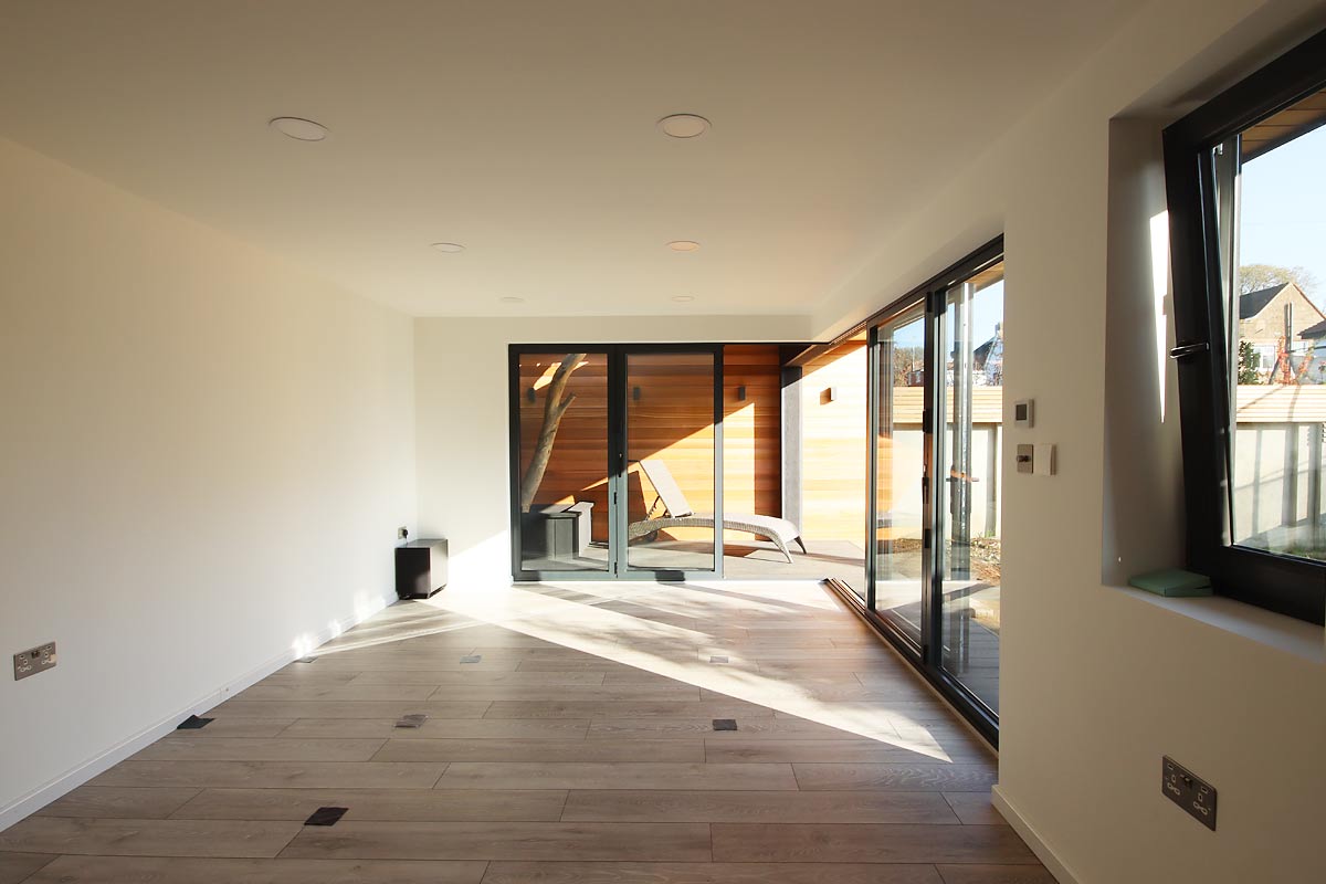 A modern, unfurnished room with light wood flooring and large sliding glass doors leading to a patio with lounge chairs. This space resembles insulated garden rooms, featuring recessed ceiling lights, white walls, a small black speaker in the corner, and several electrical outlets on the walls. - a room in the garden