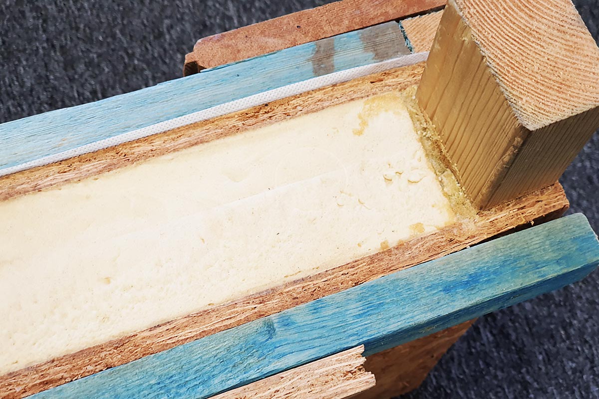 A close-up view of a partially split wooden beam showing its core. The outer layers appear to be treated wood, with one side painted light blue. The inner section is a light-colored, foam-like material, similar to those used in insulated garden rooms. The beam is placed on a dark carpeted surface. - a room in the garden