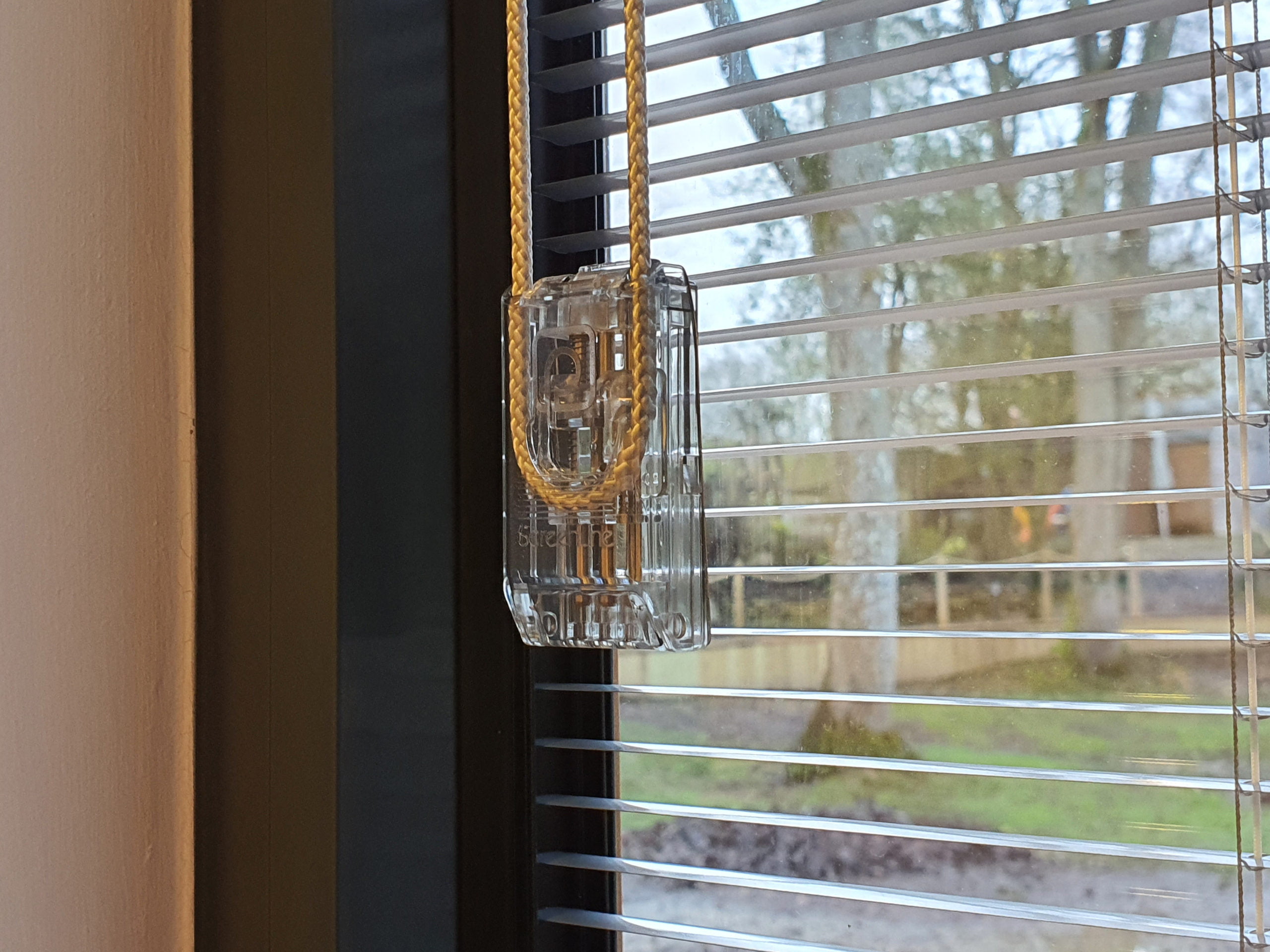 A clear plastic keycard holder with a yellow lanyard hangs from a hook in front of a window featuring integrated blinds. Outside the window, trees and a street can be seen blurred in the background. - a room in the garden