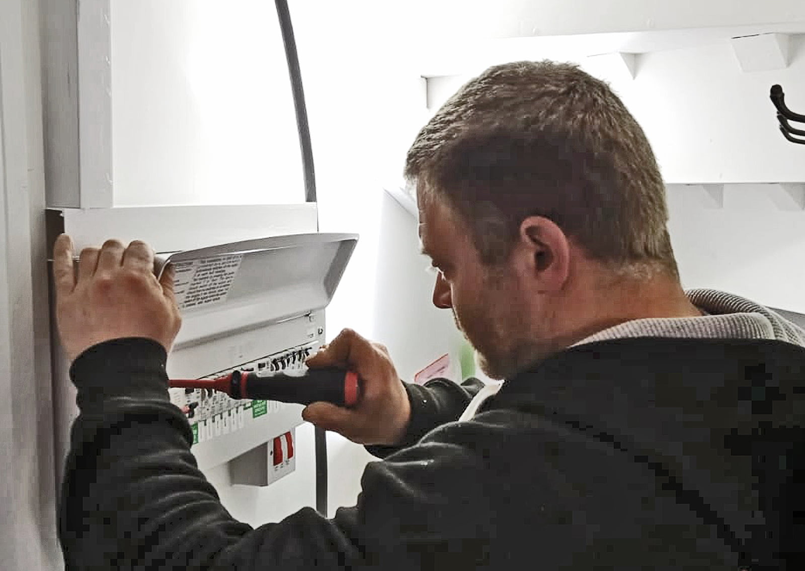 A person with short hair and a beard, dressed in a black hoodie, is using a red and black screwdriver to adjust or fix something in an electrical panel on the white wall of a bright garden room. The environment appears to be indoors with plenty of natural light. - a room in the garden