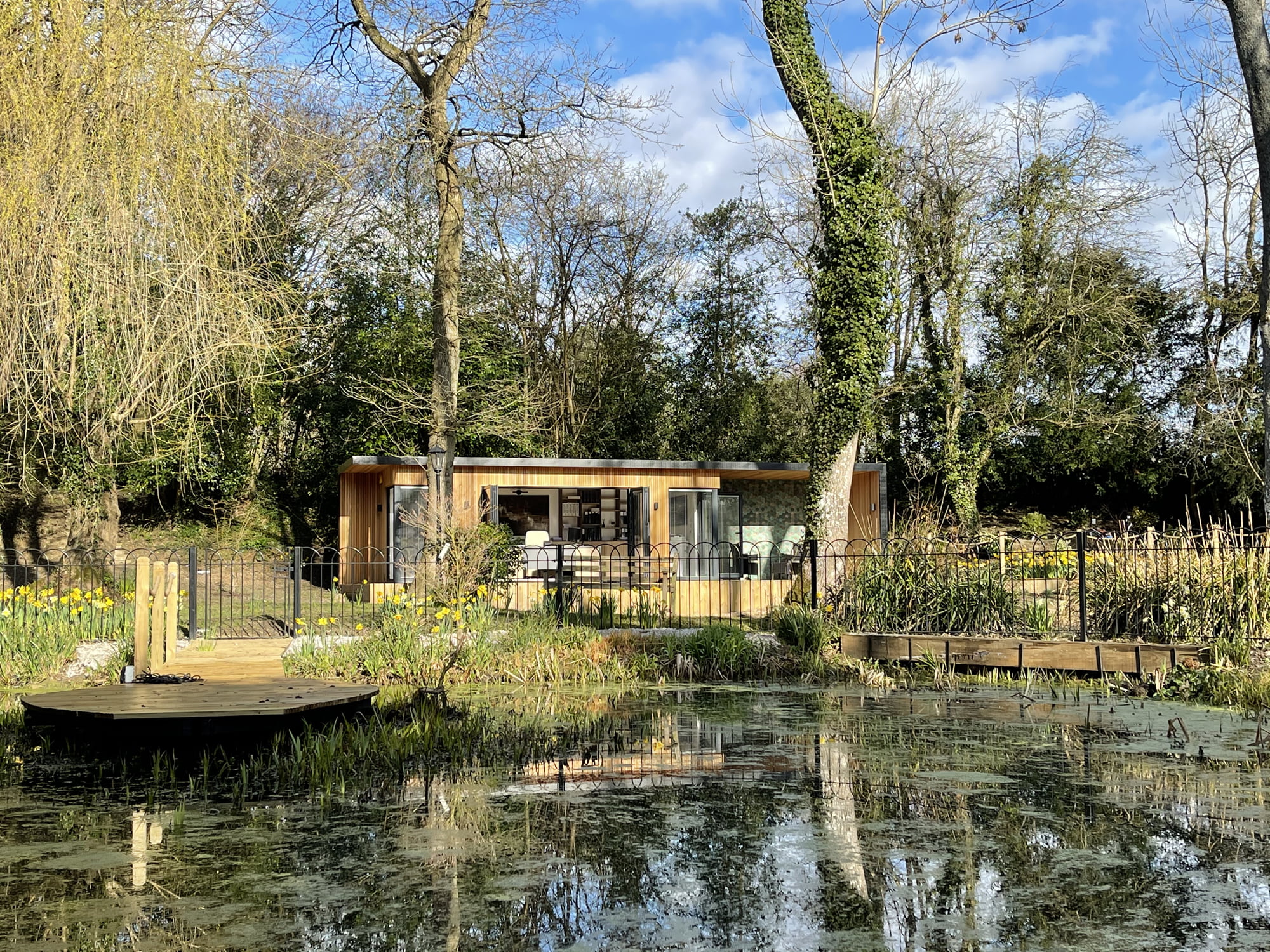 A chic, modern wooden tiny house with large glass windows stands in front of a tranquil pond surrounded by greenery and trees. Yellow flowers bloom beside the pond, adding a touch of color to the peaceful and natural setting—making it an ideal garden office by the pond. - a room in the garden