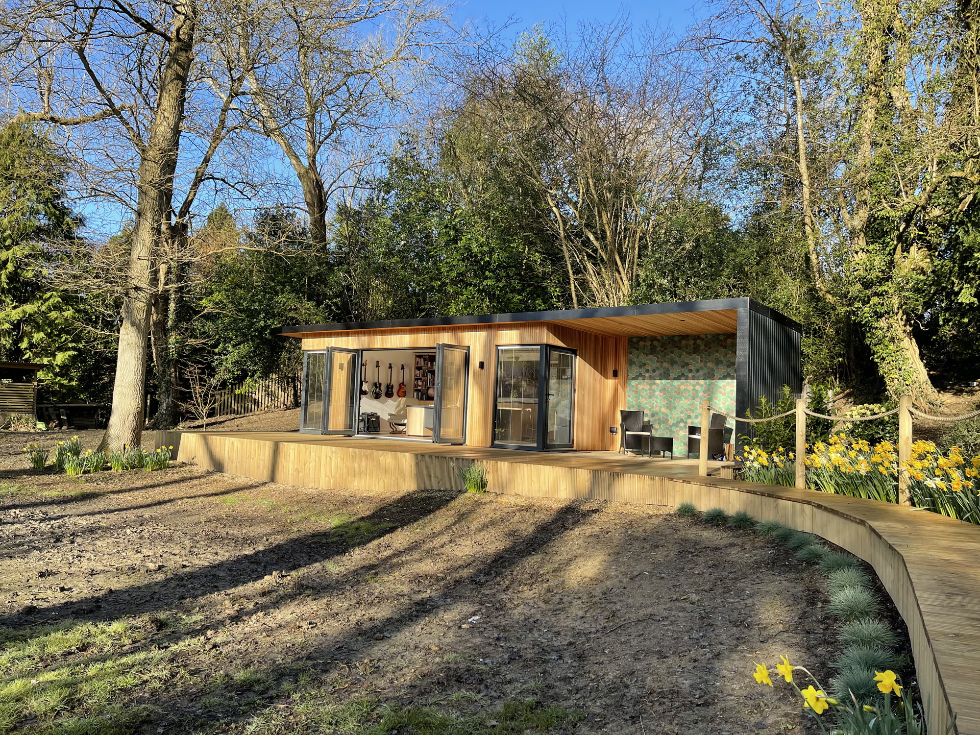 A modern, single-story wooden cabin with large glass doors sits in a woodland area on a sunny day. The cabin features a covered porch with outdoor seating, creating an ideal garden office by the pond. The surrounding yard includes blooming daffodils and trees with budding leaves. - a room in the garden