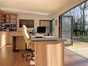 A well-lit garden office by the pond features a wooden desk, beige office chair, computer monitor, and paperwork on the desk. Shelves with books and files are on the left. Glass doors are open, leading to a deck with a view of trees outside. A pair of shoes are under the desk. - a room in the garden