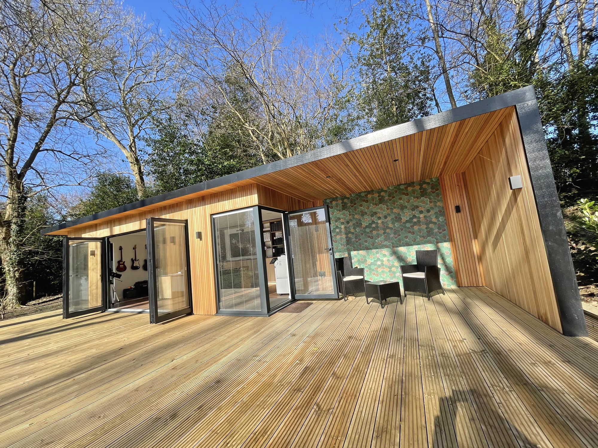 A modern wooden cabin with large glass doors and a spacious deck, perfect as a garden office by the pond. The interior, visible through the doors, includes hanging guitars and a kitchenette. The exterior features a dining table with chairs under a partially covered area. Trees surround the cabin. - a room in the garden