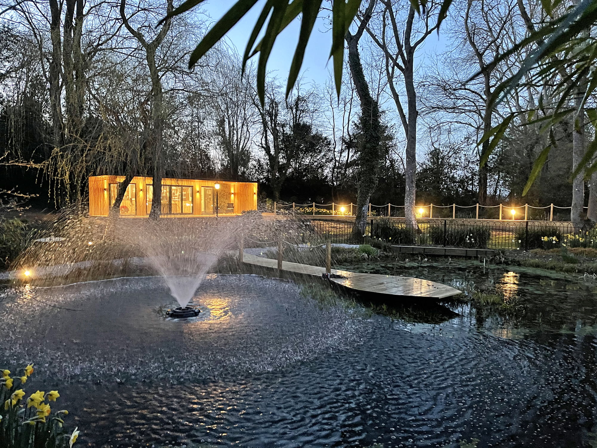 A serene garden landscape at twilight features a circular water fountain in the foreground. A wooden boardwalk extends over a pond surrounded by trees with budding leaves. In the background, a well-lit modern wooden garden office glows, and string lights outline a pathway. - a room in the garden