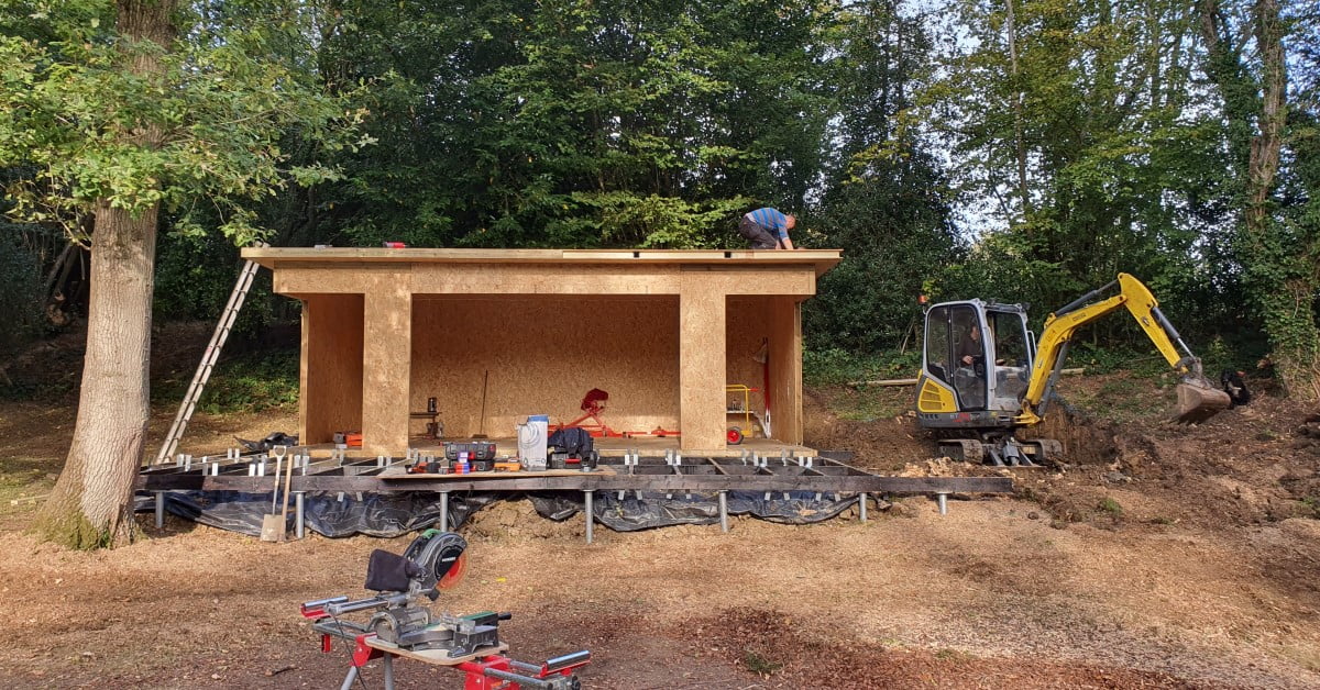 A small wooden shed is under construction near a forest. One person is working on the roof, while another sits inside the shed. A mini-excavator, its bucket digging into the ground, stands to the right. Various cost-effective construction tools are scattered around. - a room in the garden