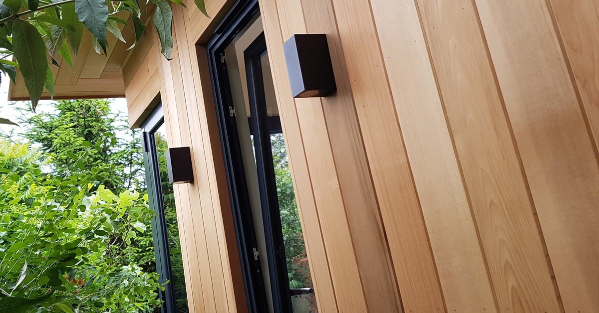 Close-up of a modern wooden house exterior featuring vertical light-colored wood panels, highlighting the cost-effective yet stylish design. Two black rectangular wall lights are mounted on the panels. Green foliage and trees surround the house, adding a natural element to the scene. - a room in the garden